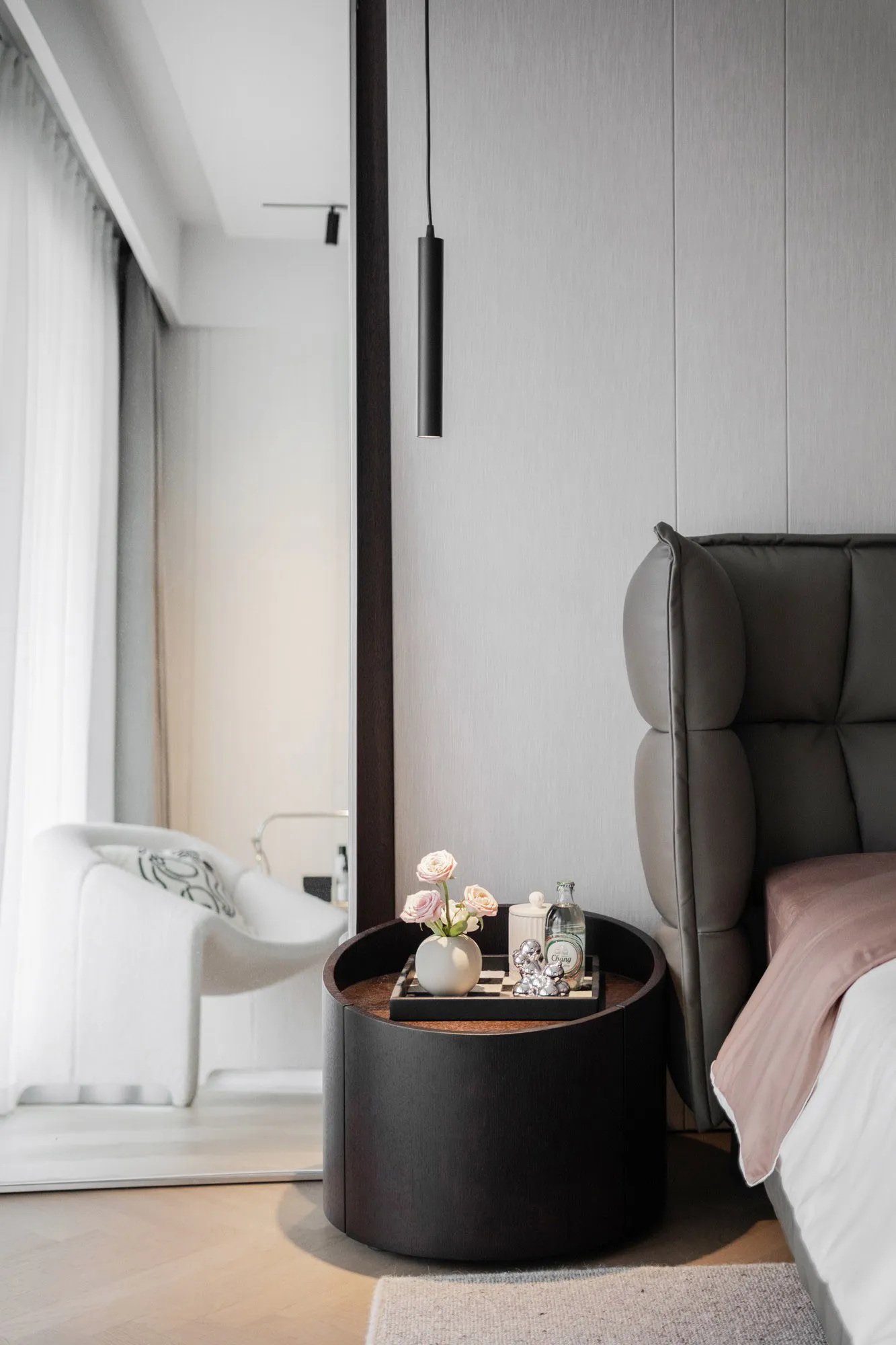 A modern minimalist bedroom featuring a textured black headboard and wooden side table with a vase of pink roses and personal
