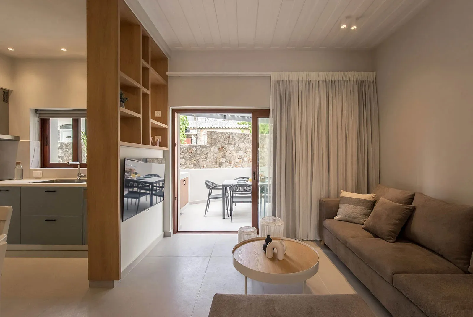 A modern, minimalist living room with concrete walls, wooden furniture, and a flat-screen TV mounted on the wall.