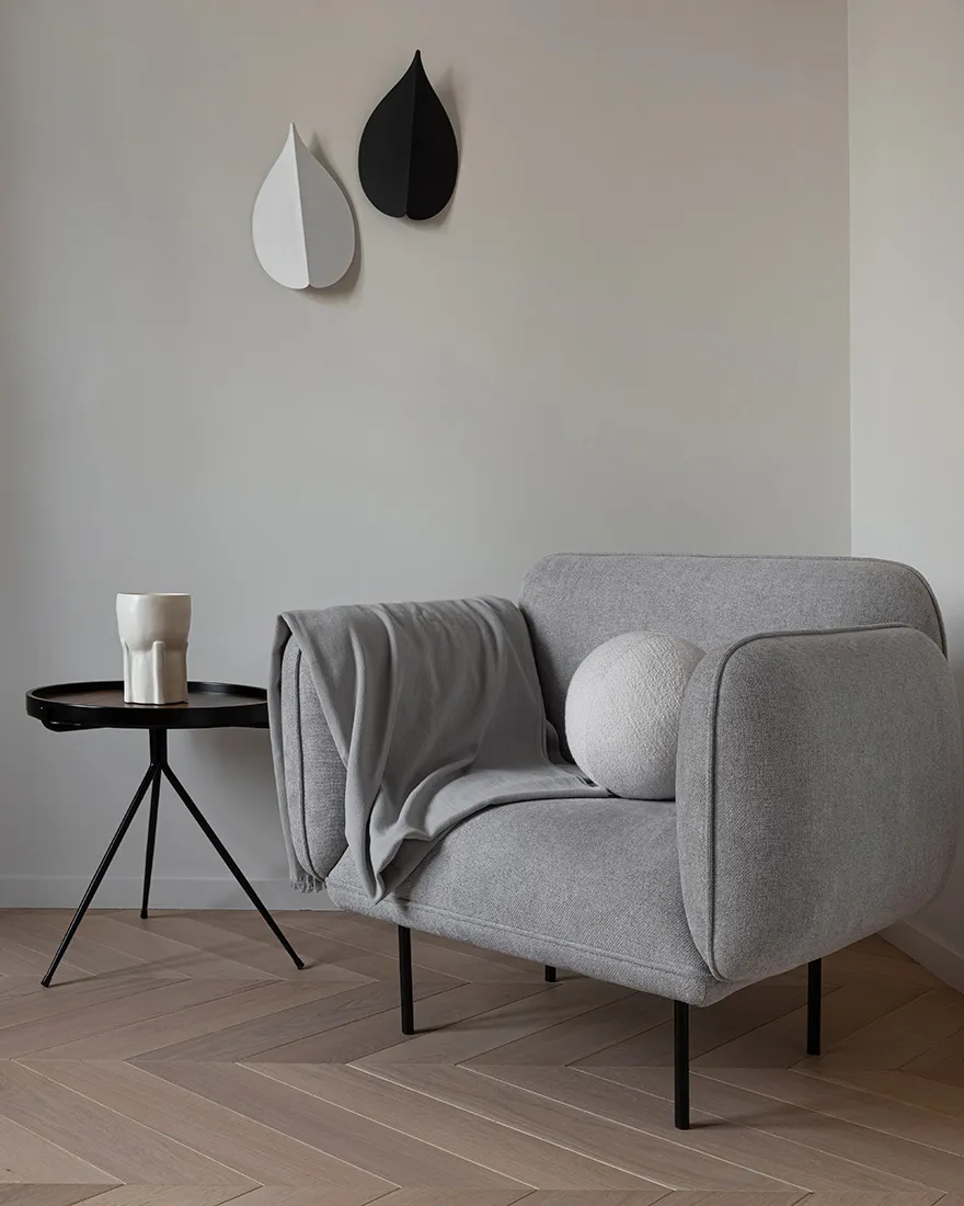 A modern minimalist living room with a grey upholstered armchair, a small round side table with a white vase, and wall art re