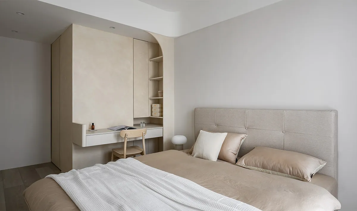 Modern, minimalist bedroom with a beige headboard bed, wooden floor, and built-in desk with shelves.