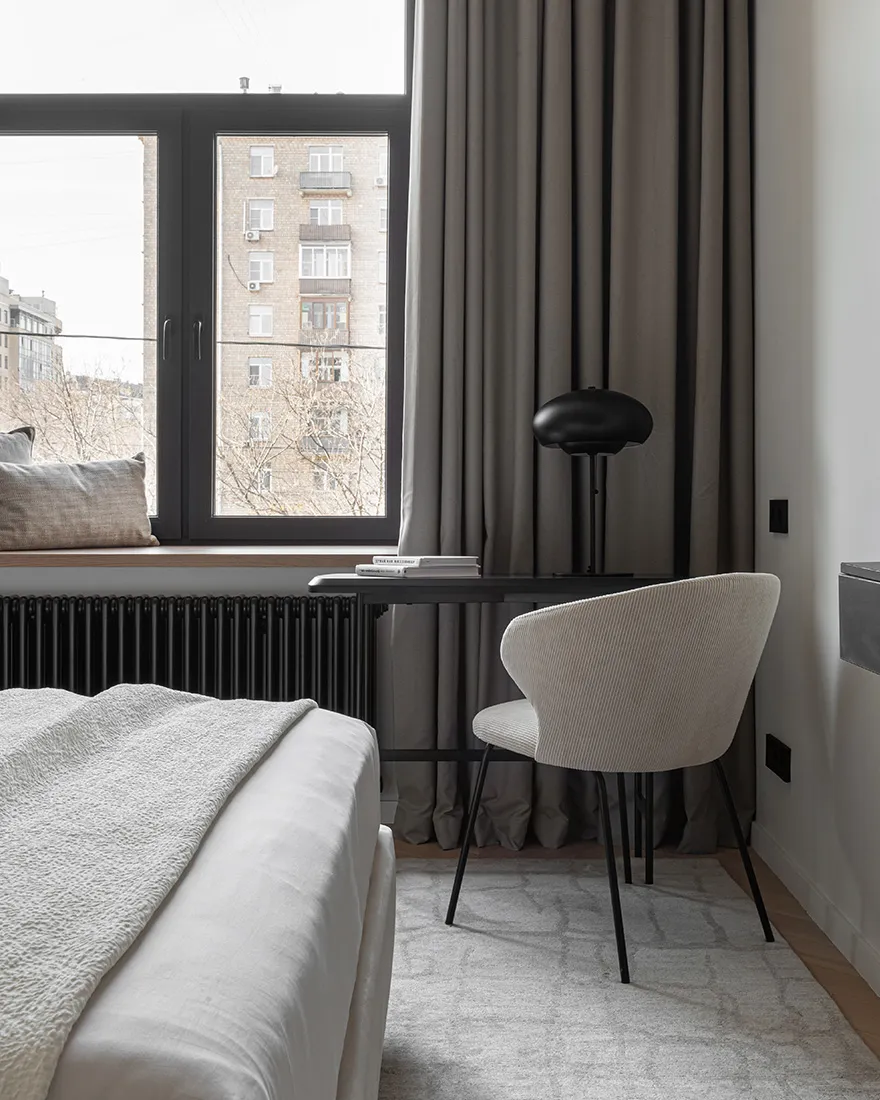 Minimalist bedroom featuring a white bed with textured fabric, a black desk, and a beige chair.
