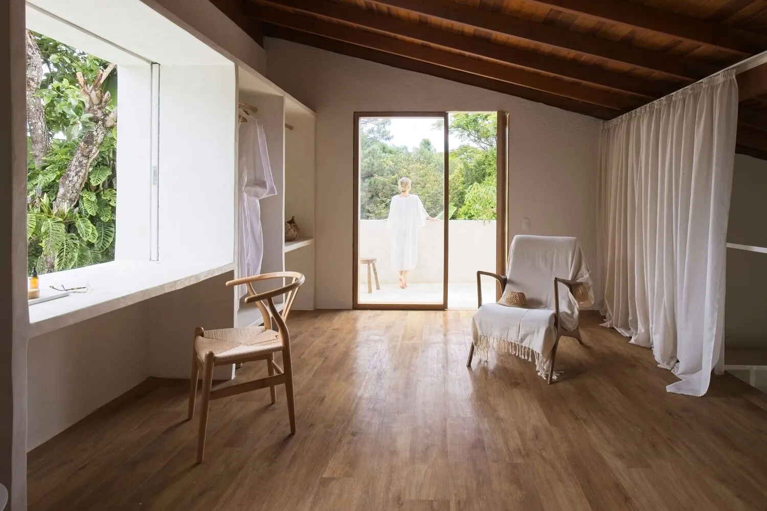 A minimalist room with wooden floors, a white chair, and a glass door leading to a balcony with greenery outside.