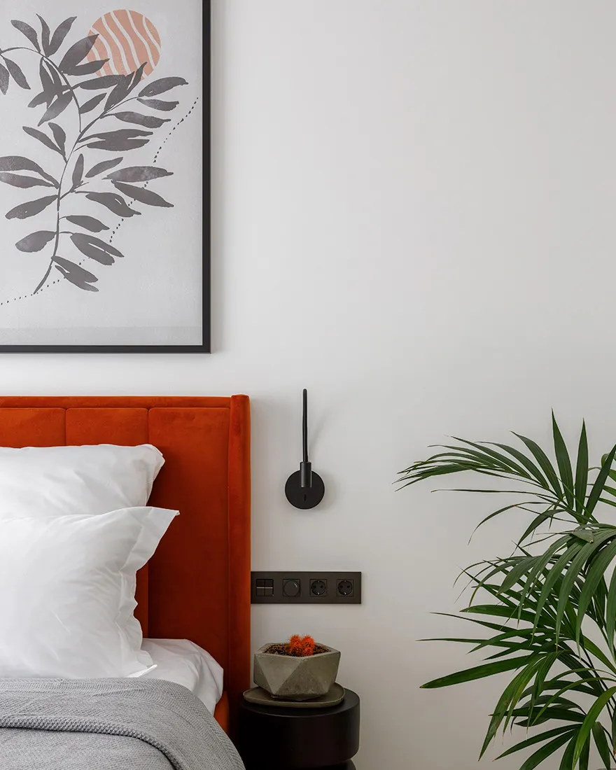 A modern bedroom featuring an orange velvet headboard, white pillows, and a grey throw blanket, complemented by a concrete be