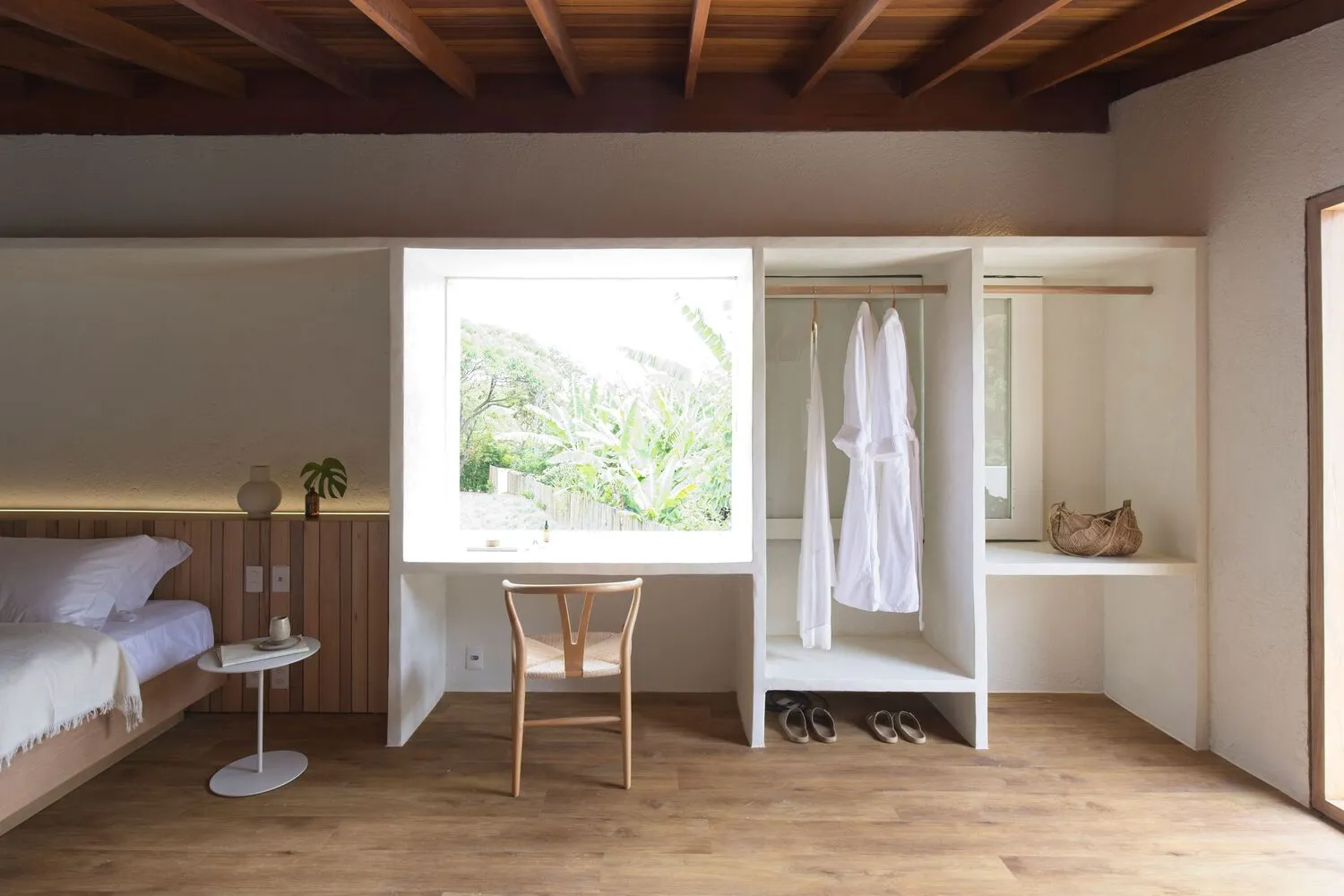 A minimalist bedroom with a large window, a wooden chair, and a view of an indoor garden.