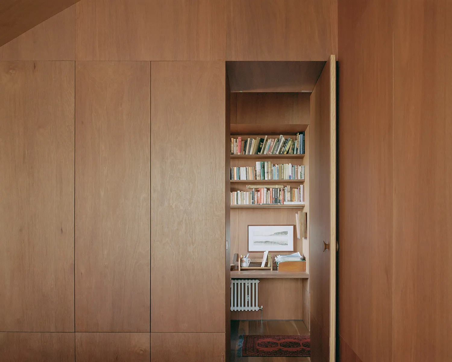 A minimalist library with wooden shelves filled with books, a desk, and a white radiator beneath a wooden slat wall.