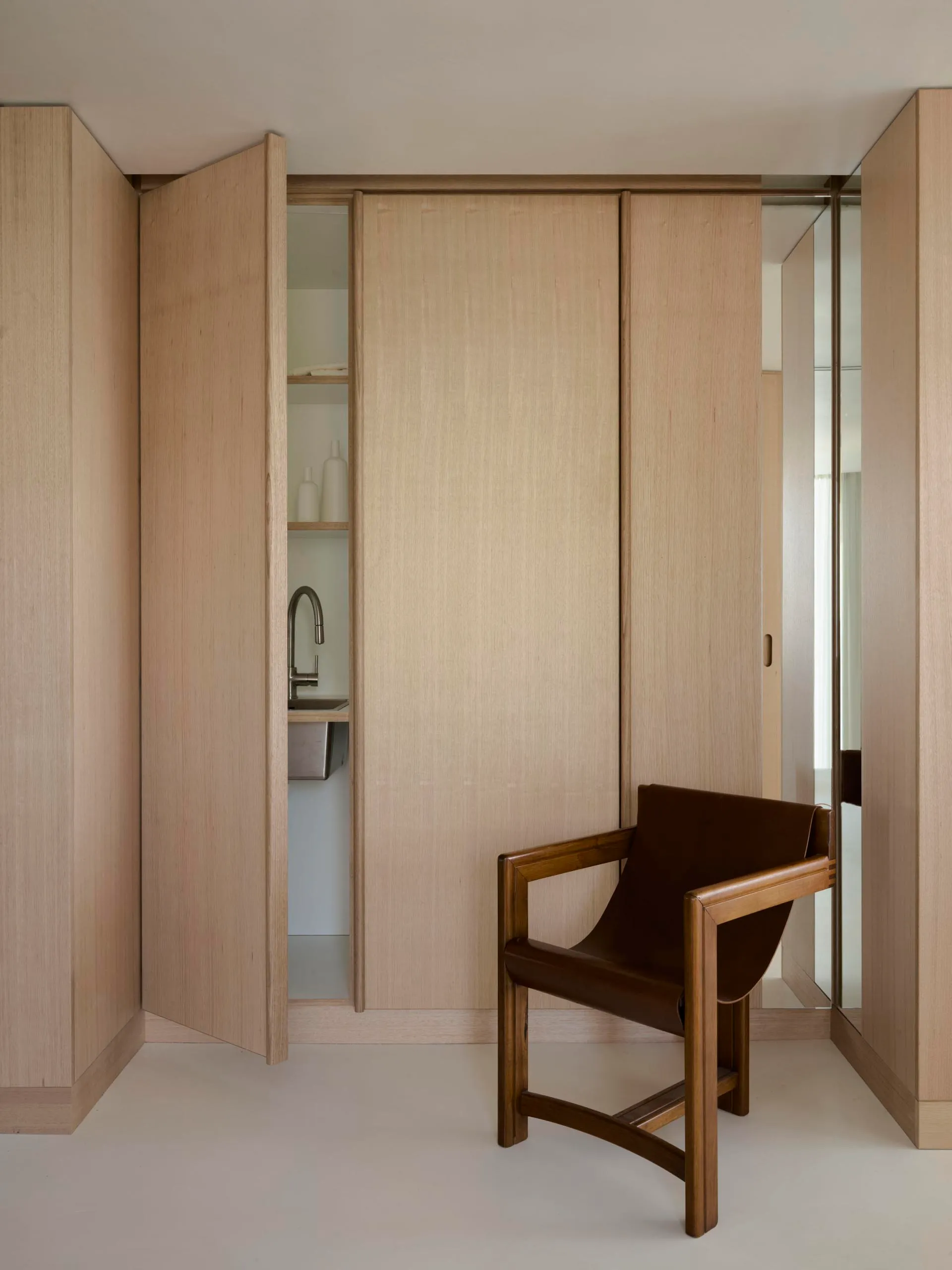 A minimalist bedroom with wooden walls and floor, featuring a chair in the foreground and a small kitchen area in the backgro