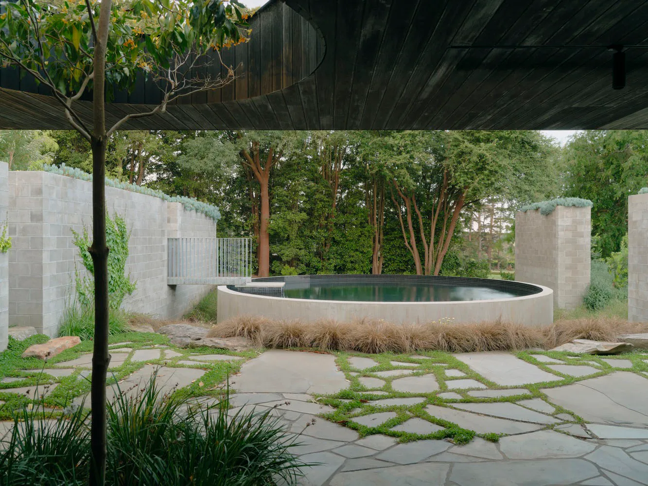 A serene outdoor space featuring a round pool surrounded by stone walls, surrounded by lush greenery and a pathway of irregul