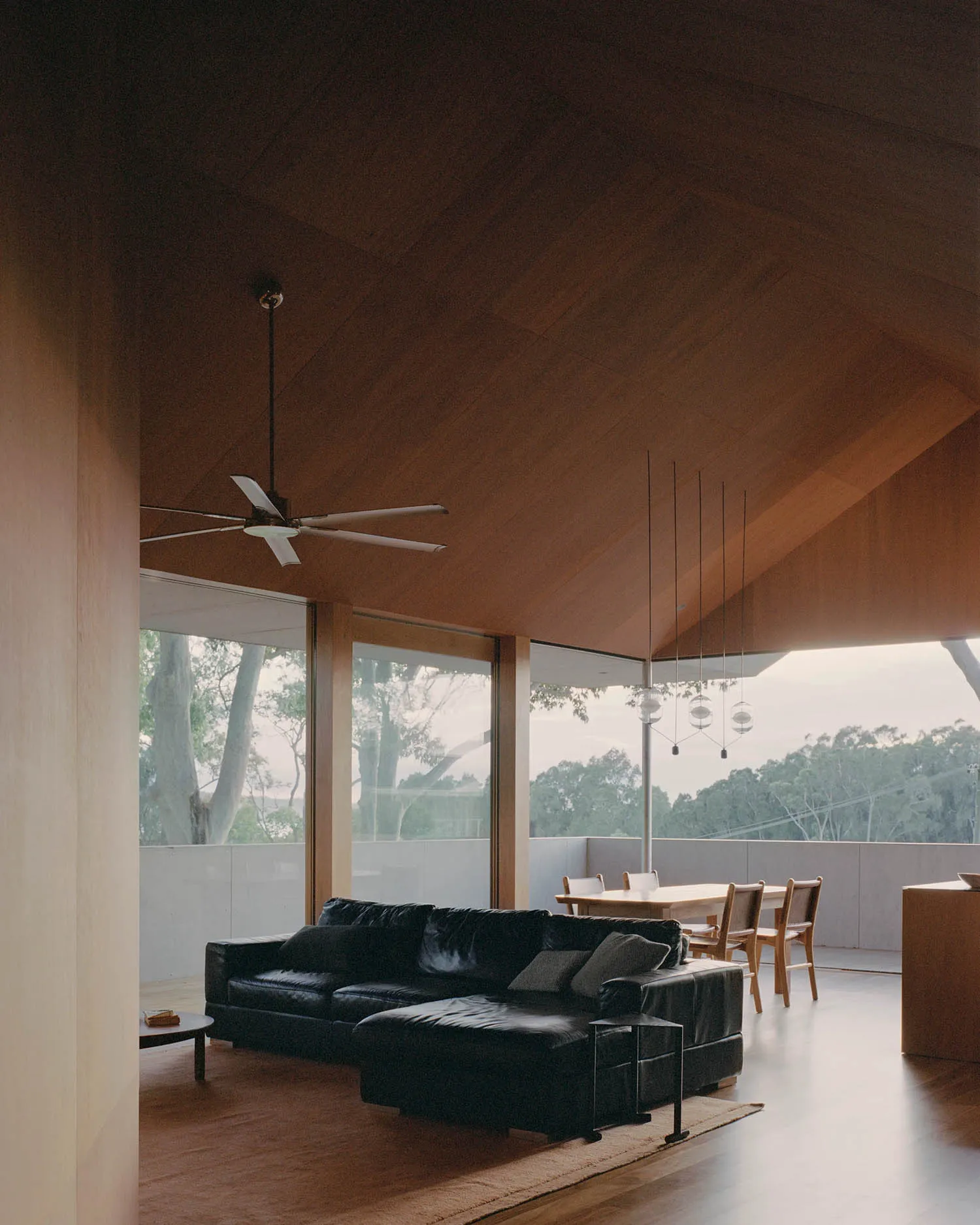 A modern living room with a black leather sofa, wooden ceiling, and large windows overlooking trees.