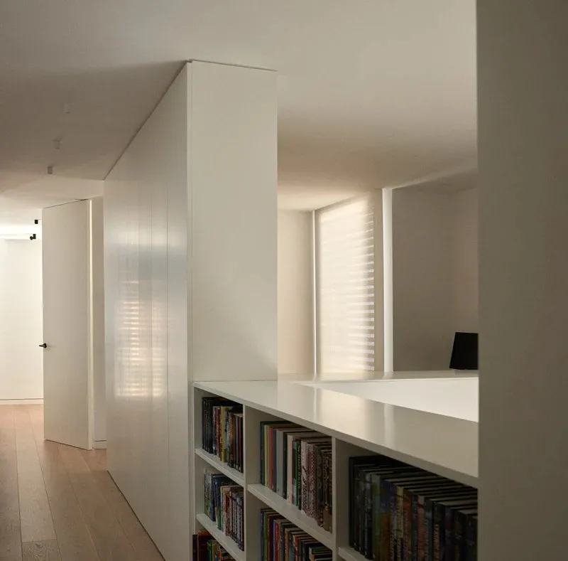 Contemporary minimalist library with white bookshelves displaying assorted books and a window with closed blinds.