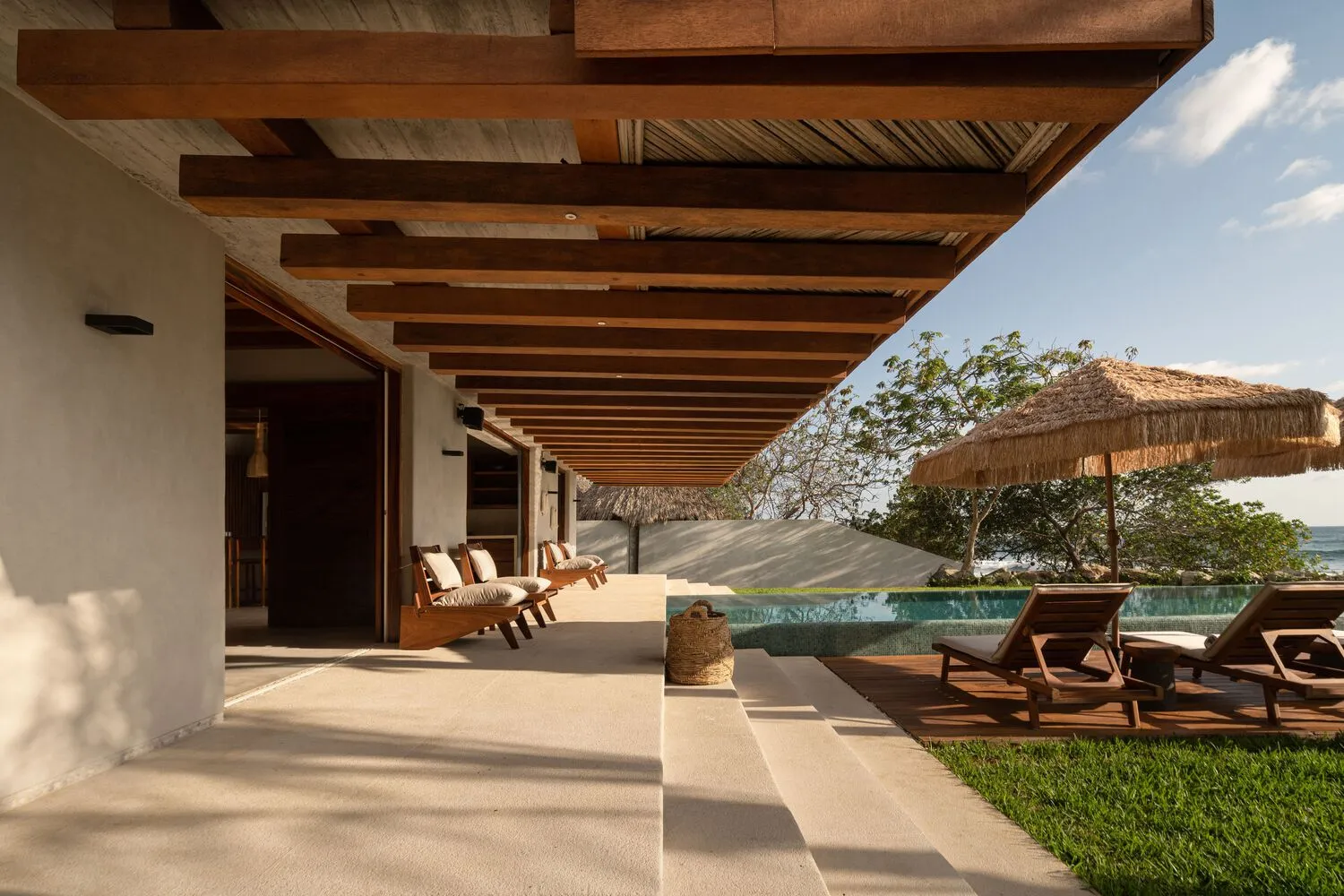 Modern minimalist residential patio with wooden furniture, thatched umbrella, and infinity pool.
