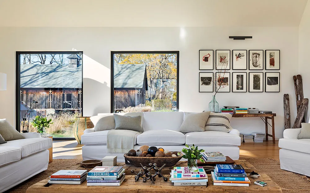 A modern minimalist living room with white furniture, wooden floors, and large windows showcasing a wooden barn outside.
