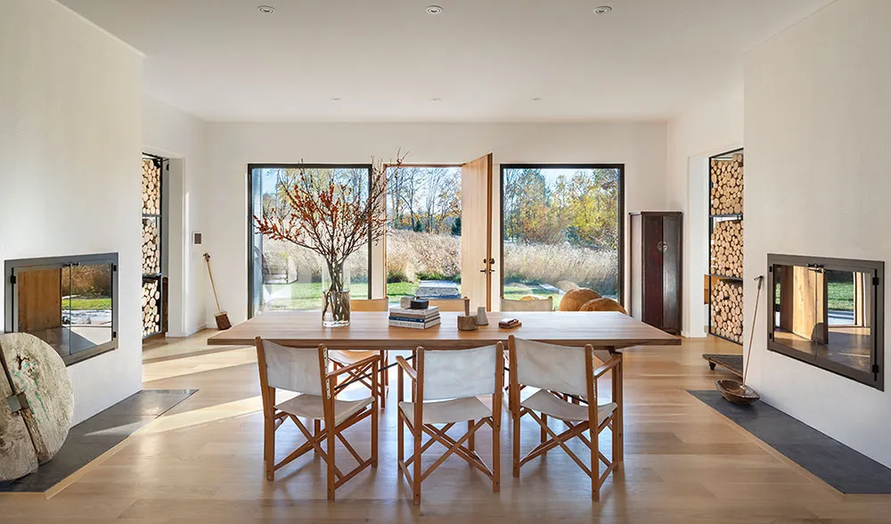A modern residential dining room with a wooden table and chairs, large windows, and firewood storage.