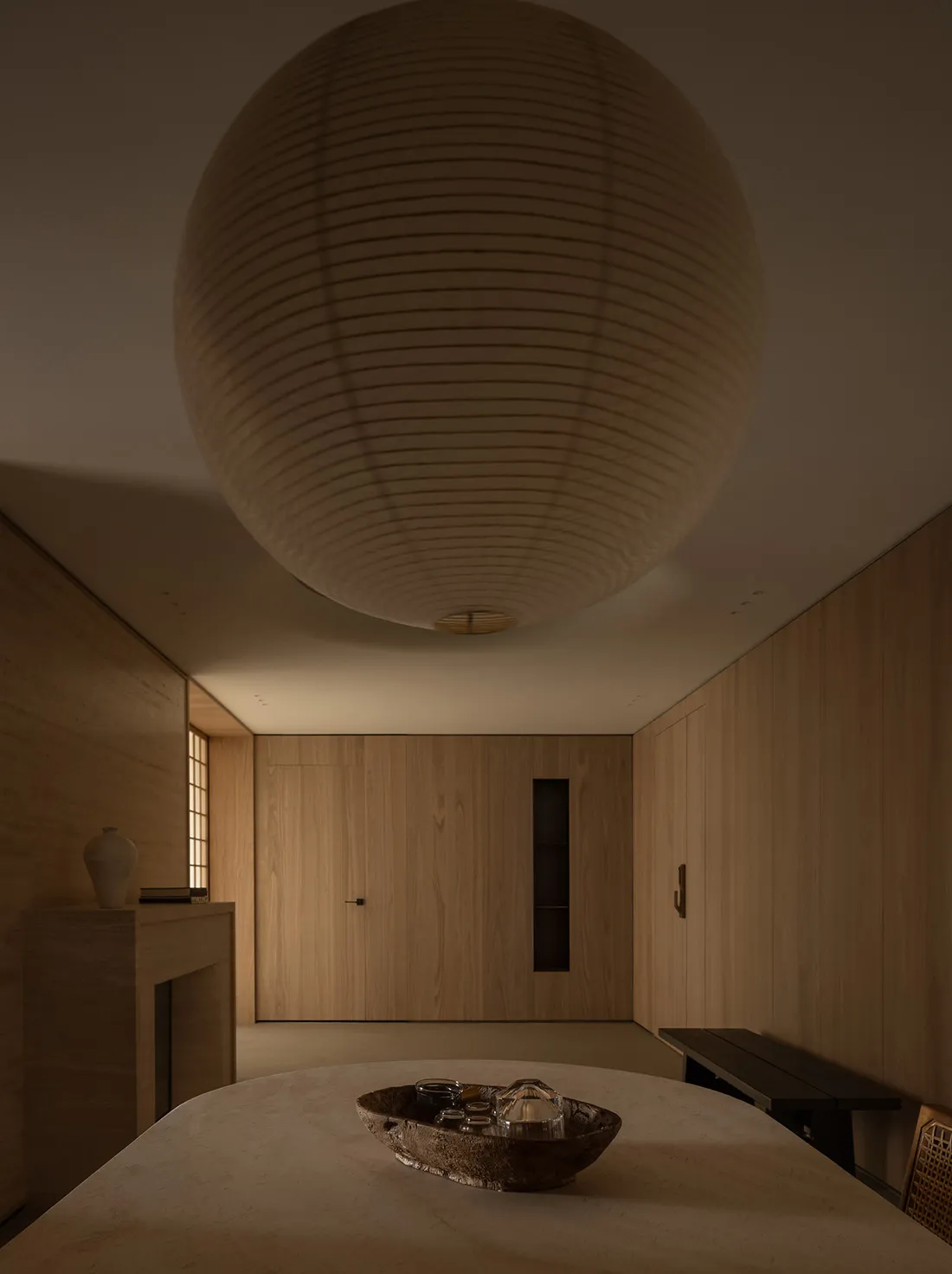 A minimalist interior space with wooden walls and furniture, featuring a large lamp and a bowl of objects on a table.