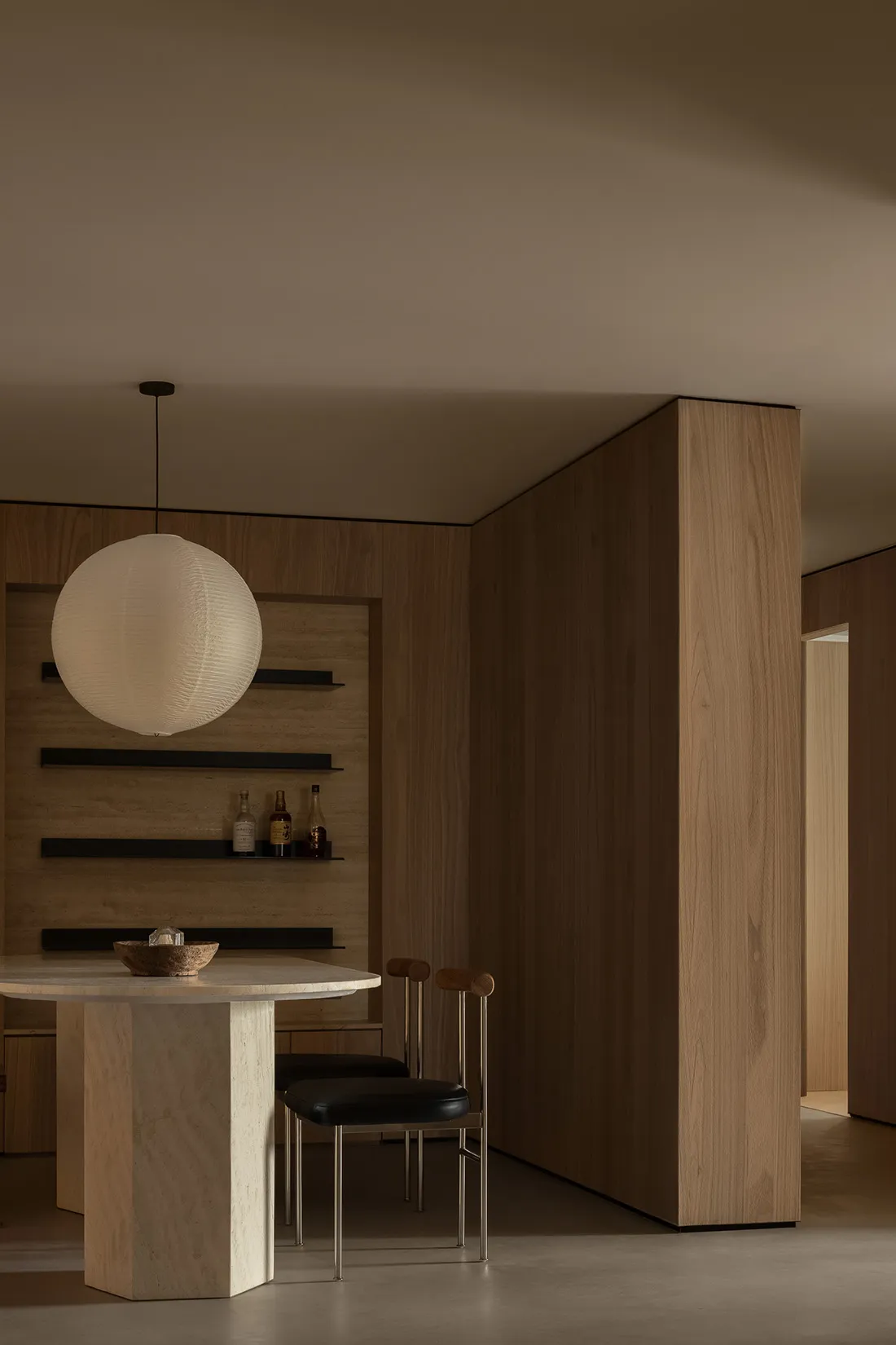 A modern dining room featuring wooden walls and chairs, with a round table, pendant light, and shelves.