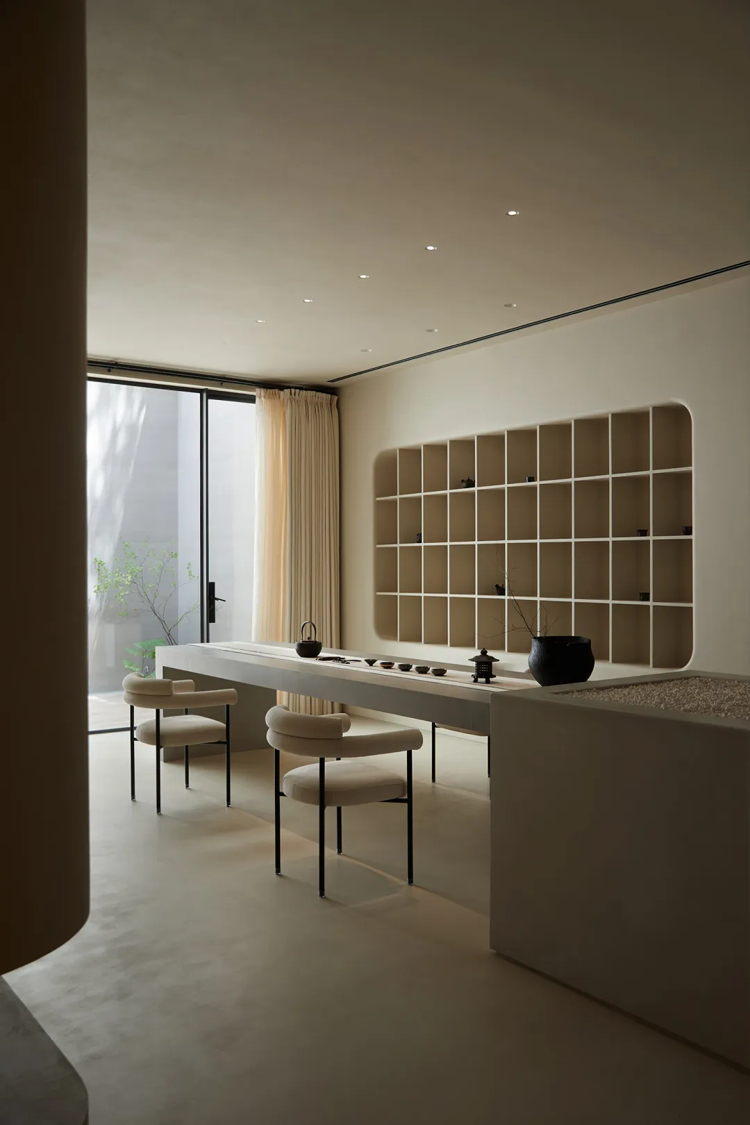 A minimalist interior space featuring concrete furniture and flooring, with a modern bookshelf and seating area by a large wi