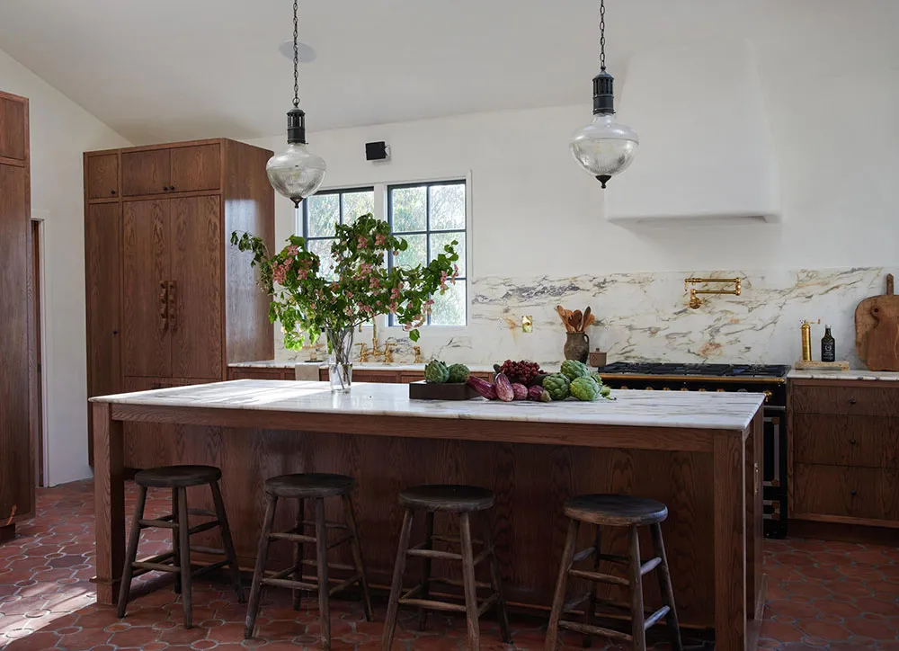 A modern, minimalist residential kitchen with a marble backsplash, wooden cabinets, and a large island with stools.