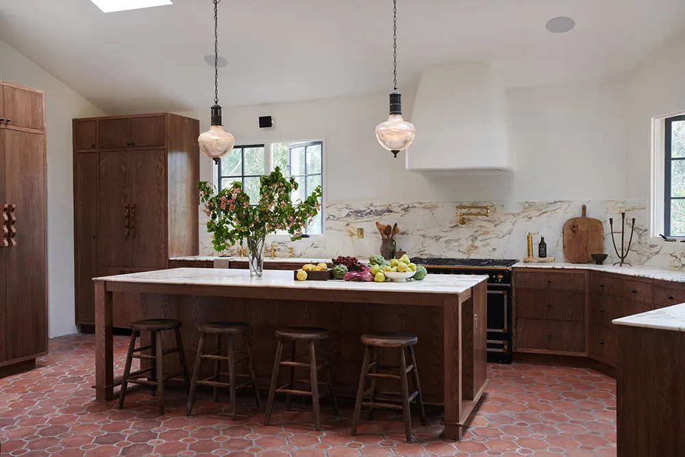 A modern kitchen with an island, wooden stools, marble countertops, and a large window.
