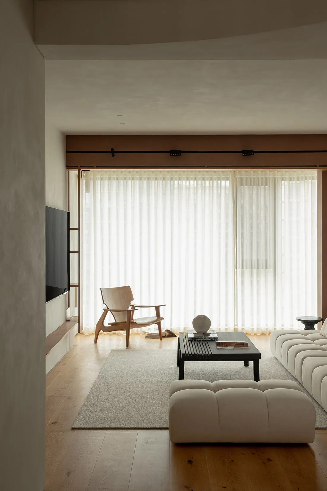 Modern minimalist living room with natural wood floor, white fabric sofa, glass coffee table, and white sheer curtains.