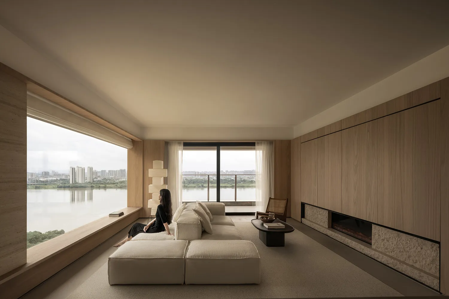 A minimalist residential living room with a large window overlooking a serene river view, featuring a beige modular sofa, a g