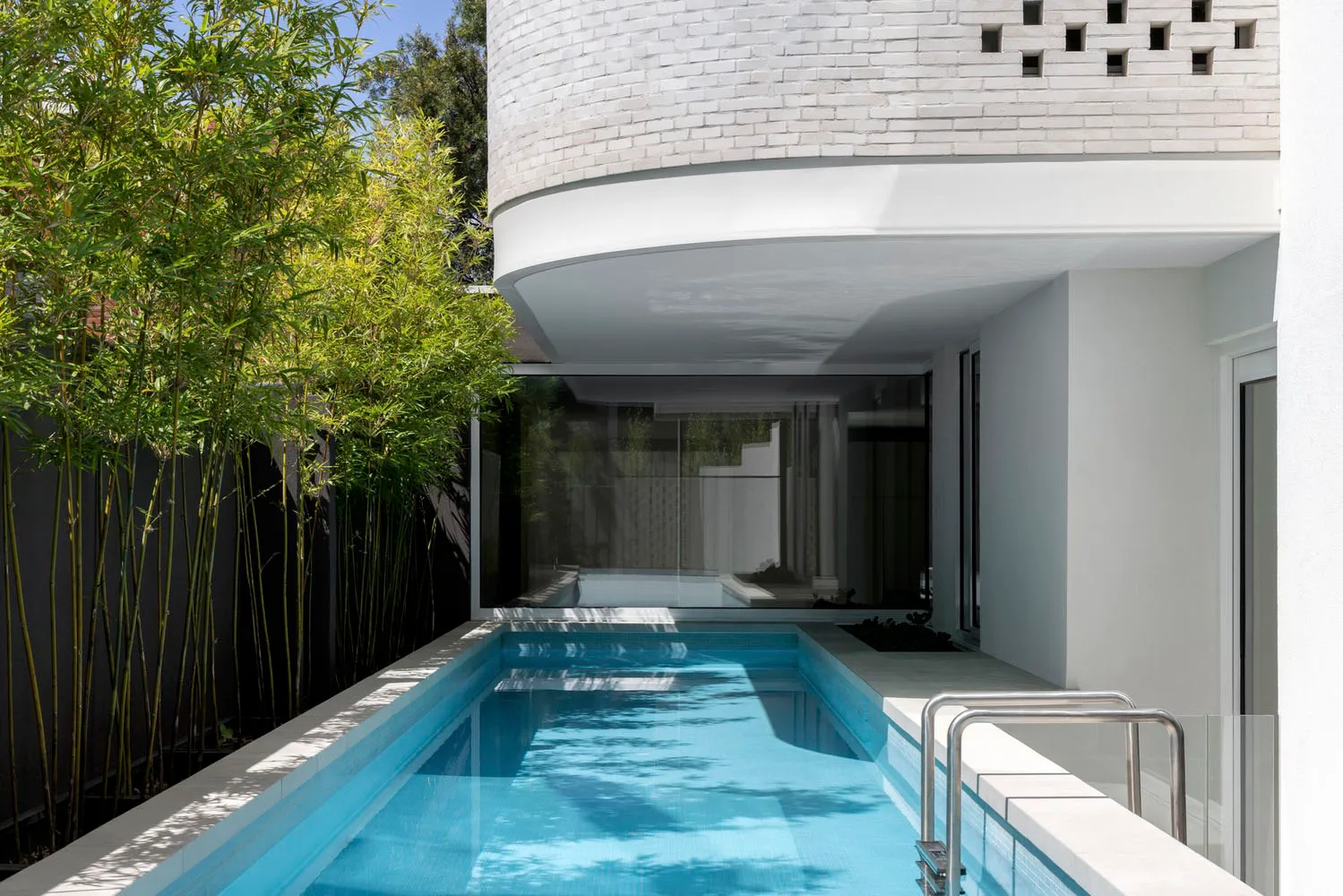 A modern house with a white brick facade and a glass swimming pool surrounded by greenery.
