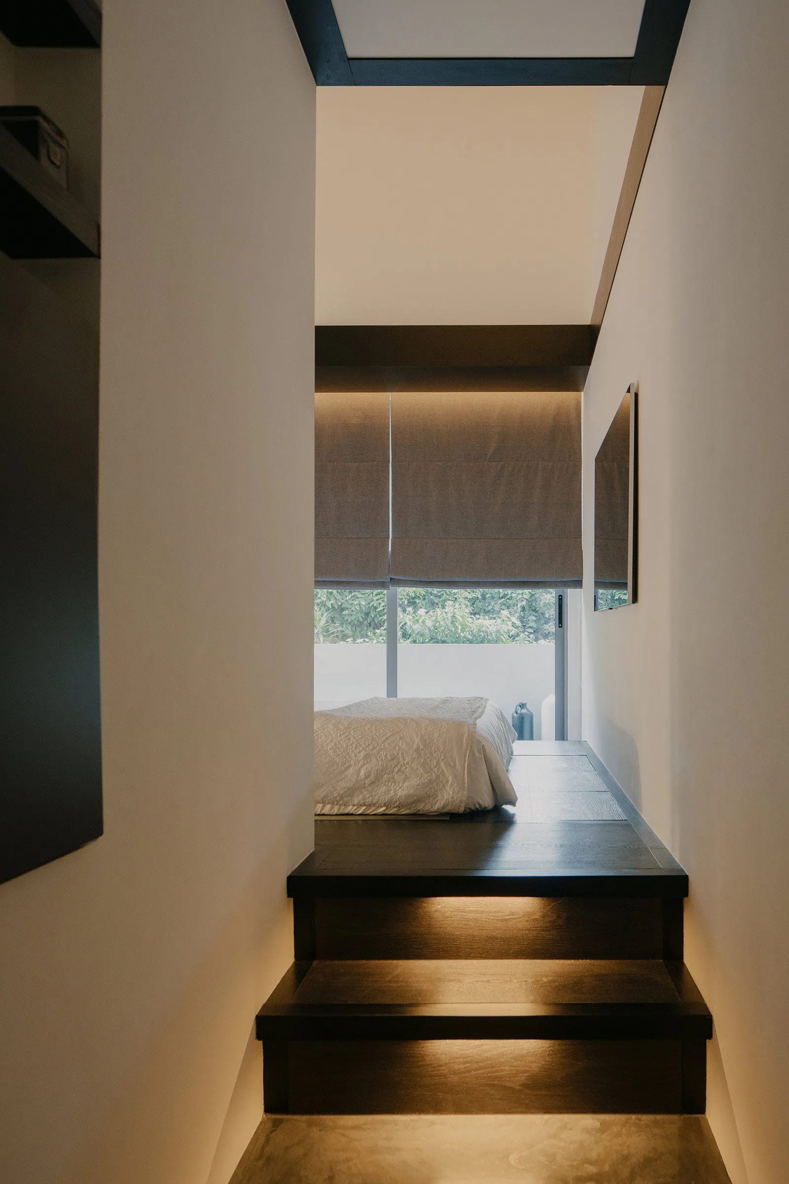 A minimalistic bedroom with a stairway leading to it, featuring a bed with white bedding, dark wood stairs with embedded ligh