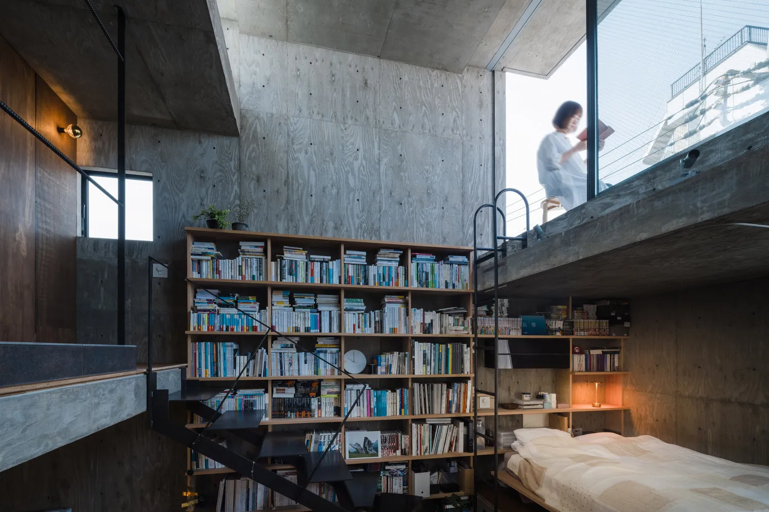 A modern industrial bedroom featuring concrete walls, a wooden bookshelf filled with books, and a black metal staircase.