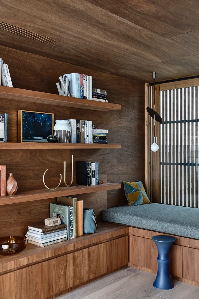 A cozy home office with wooden shelves, a cushioned bench, and a variety of books and decorative items.