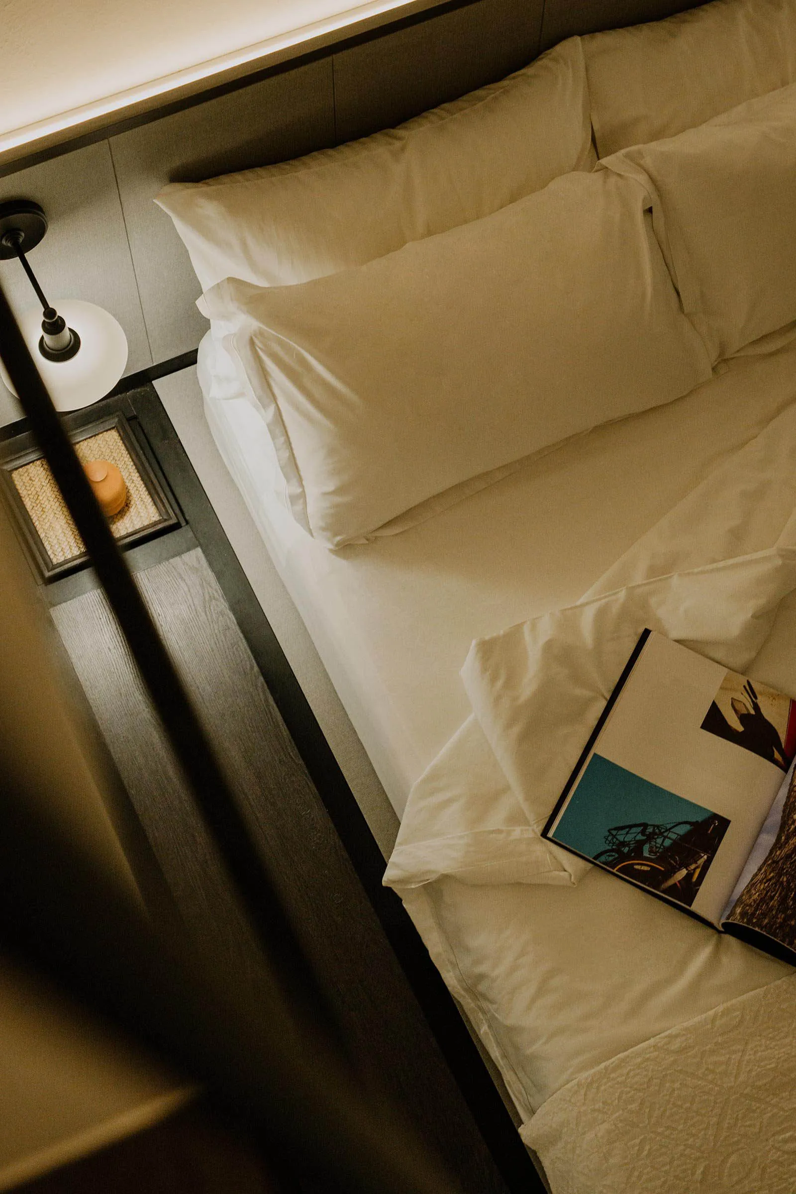 A neatly made white bed with pillows and a magazine on the mattress.