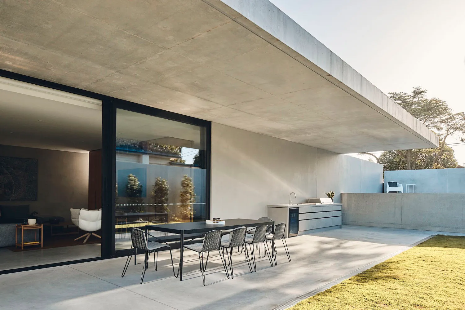 Outdoor kitchen with concrete patio, table, chairs, and modern appliances under a sloping concrete roof.