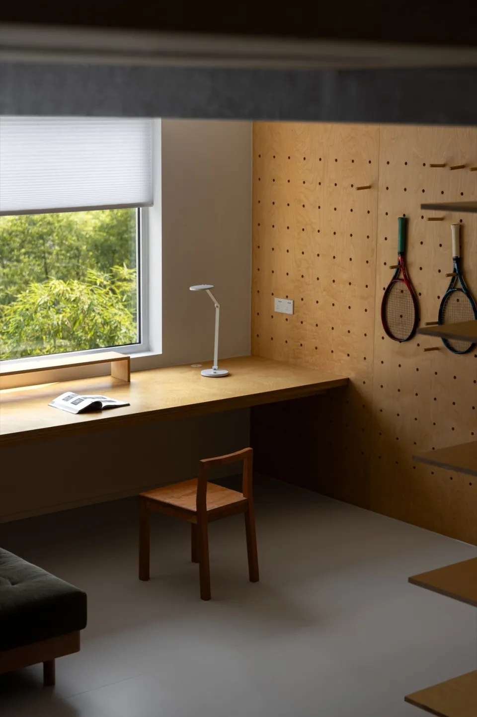 A minimalist workspace featuring a wooden desk with a lamp and a chair, mounted shelves, and two tennis rackets hanging on a