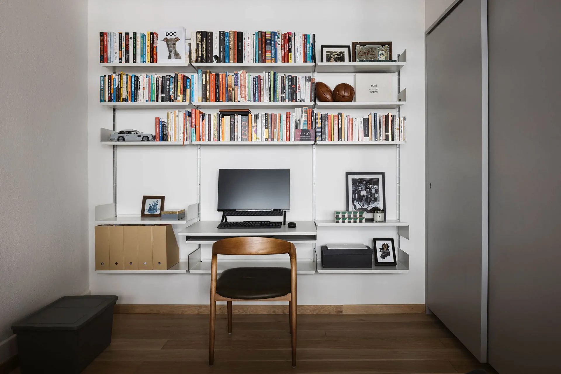 A modern home office featuring a desk with a computer setup and a wall-mounted shelving system filled with books and decorati