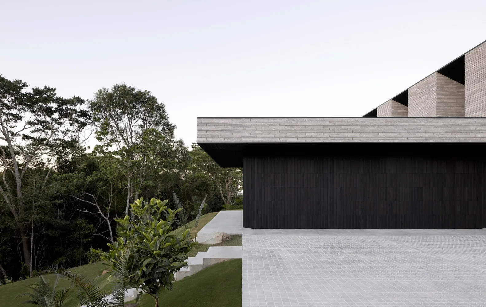 A modern residential home with a concrete exterior and a large driveway leading up to it, surrounded by lush greenery and tre