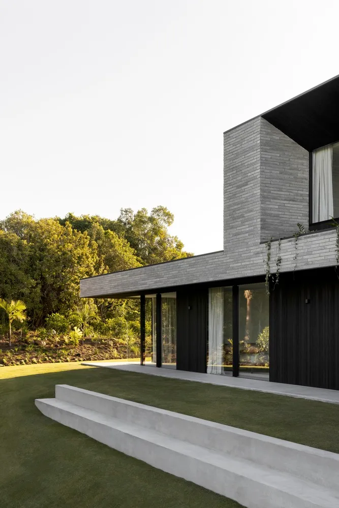 Modern residential exterior featuring concrete and glass materials, with a lush green garden and natural lighting.