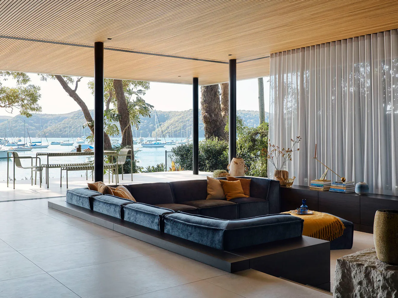 A minimalist living room with a blue sectional sofa, wooden flooring, and large windows overlooking a waterfront view.