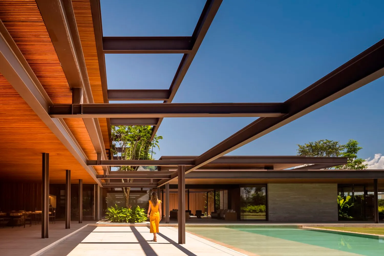 A modern architectural structure featuring a wooden roof supported by metal beams and a pool with greenery in the background.