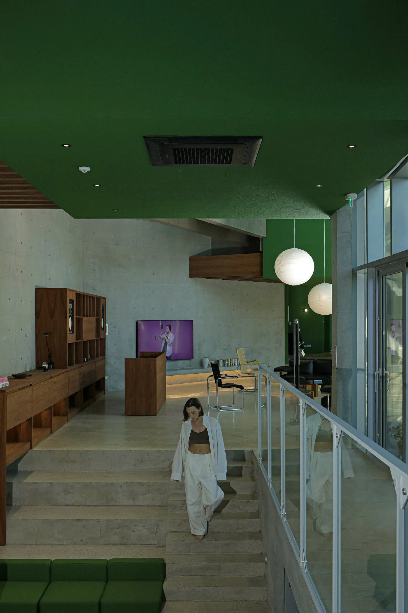 A modern living room with concrete cladding and green accent wall, where a woman walks up the stairs.