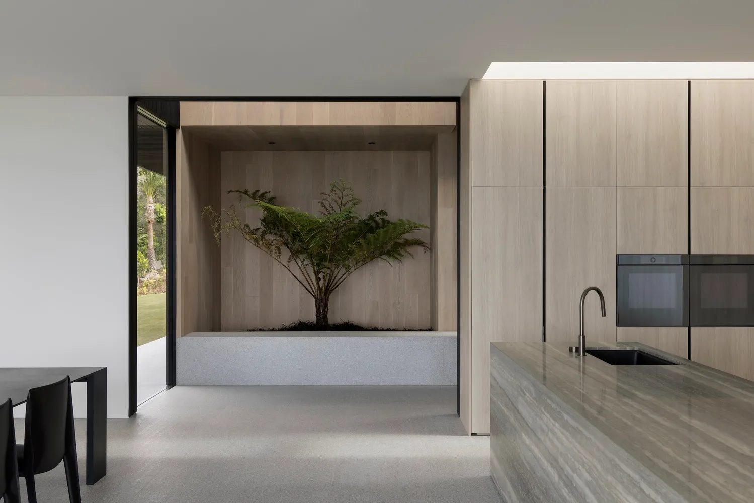 A modern residential kitchen featuring a tree embedded in a concrete planter, surrounded by wood cabinets and a marble counte
