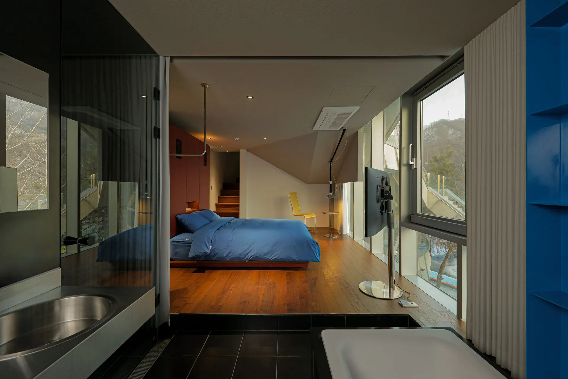 Modern bedroom featuring wood flooring and furniture, with large windows and a minimalist design.