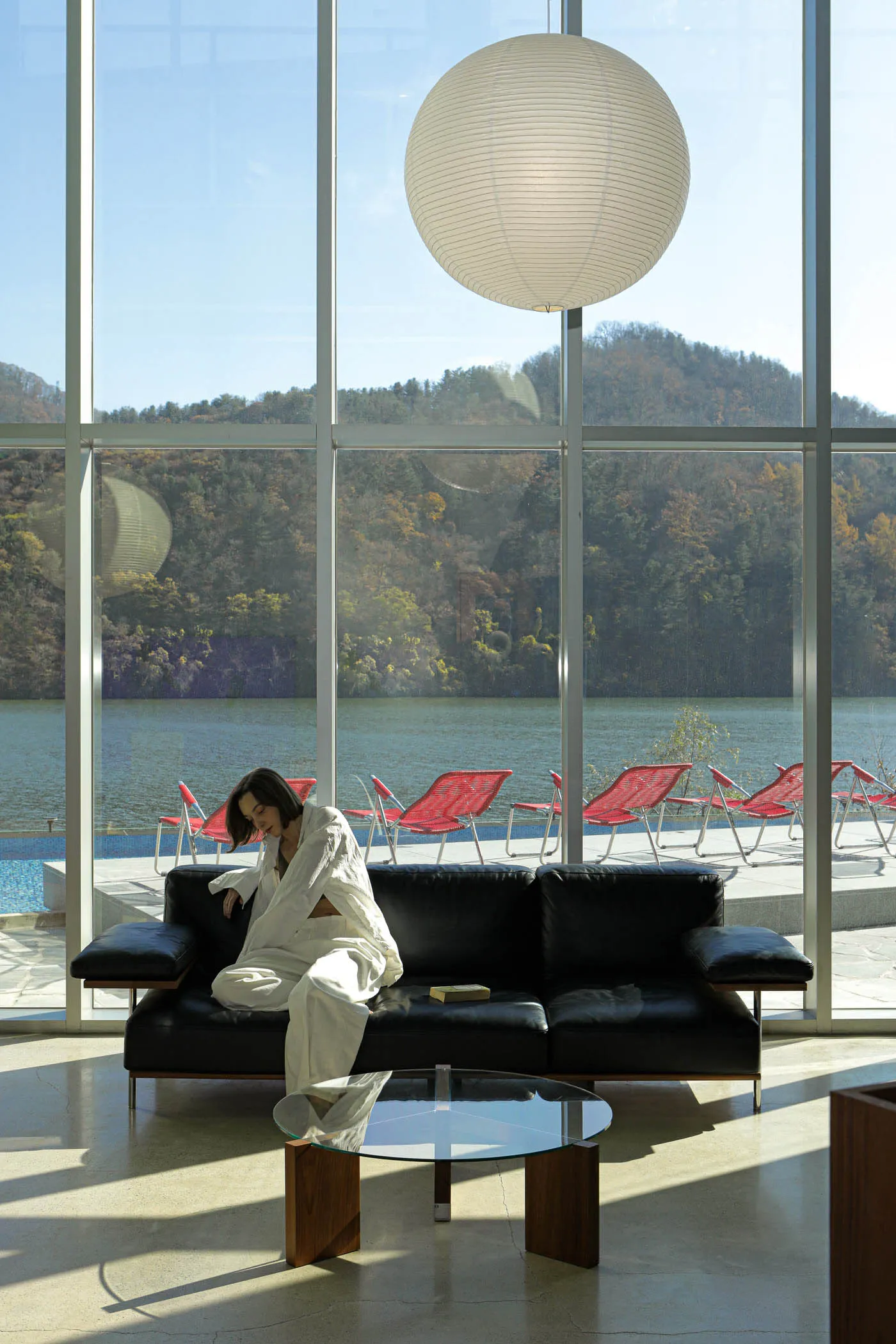 A modern interior featuring a glass window frame, with a black sofa, red lounge chairs, and a large white light fixture.
