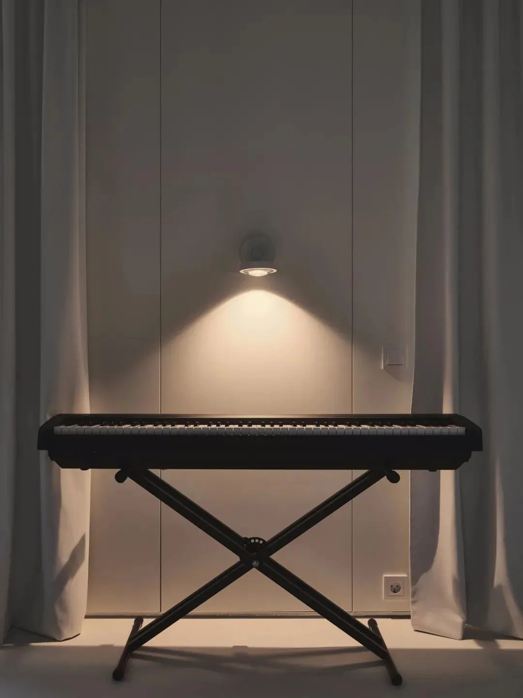A minimalist music room featuring a black electronic keyboard on a stand, backlit by a wall-mounted light.