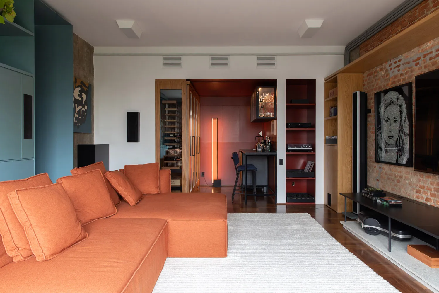 A modern living room featuring an orange sectional couch, brick walls, and wooden shelving.