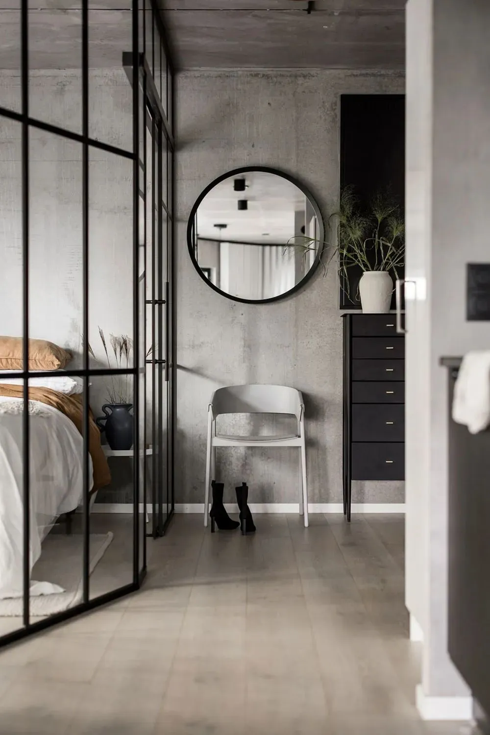 A minimalist bedroom with concrete walls, a glass partition, and a round mirror.