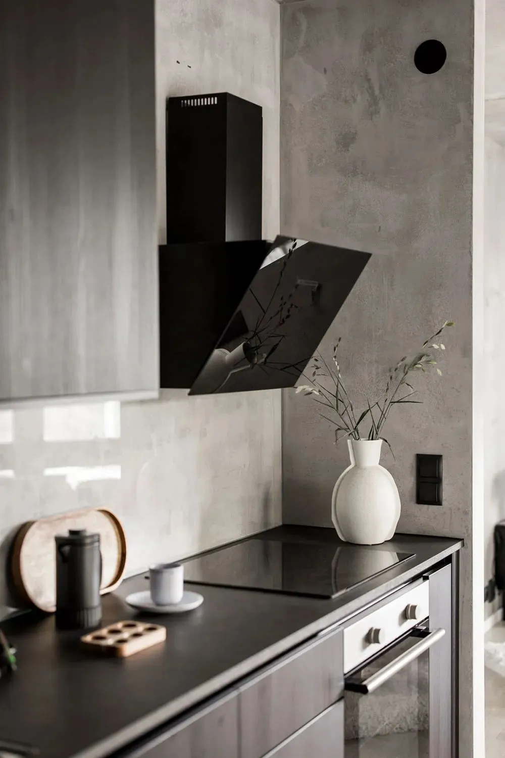 Modern kitchen with concrete finishes, featuring a sleek black stove top and a minimalist vase with greenery.