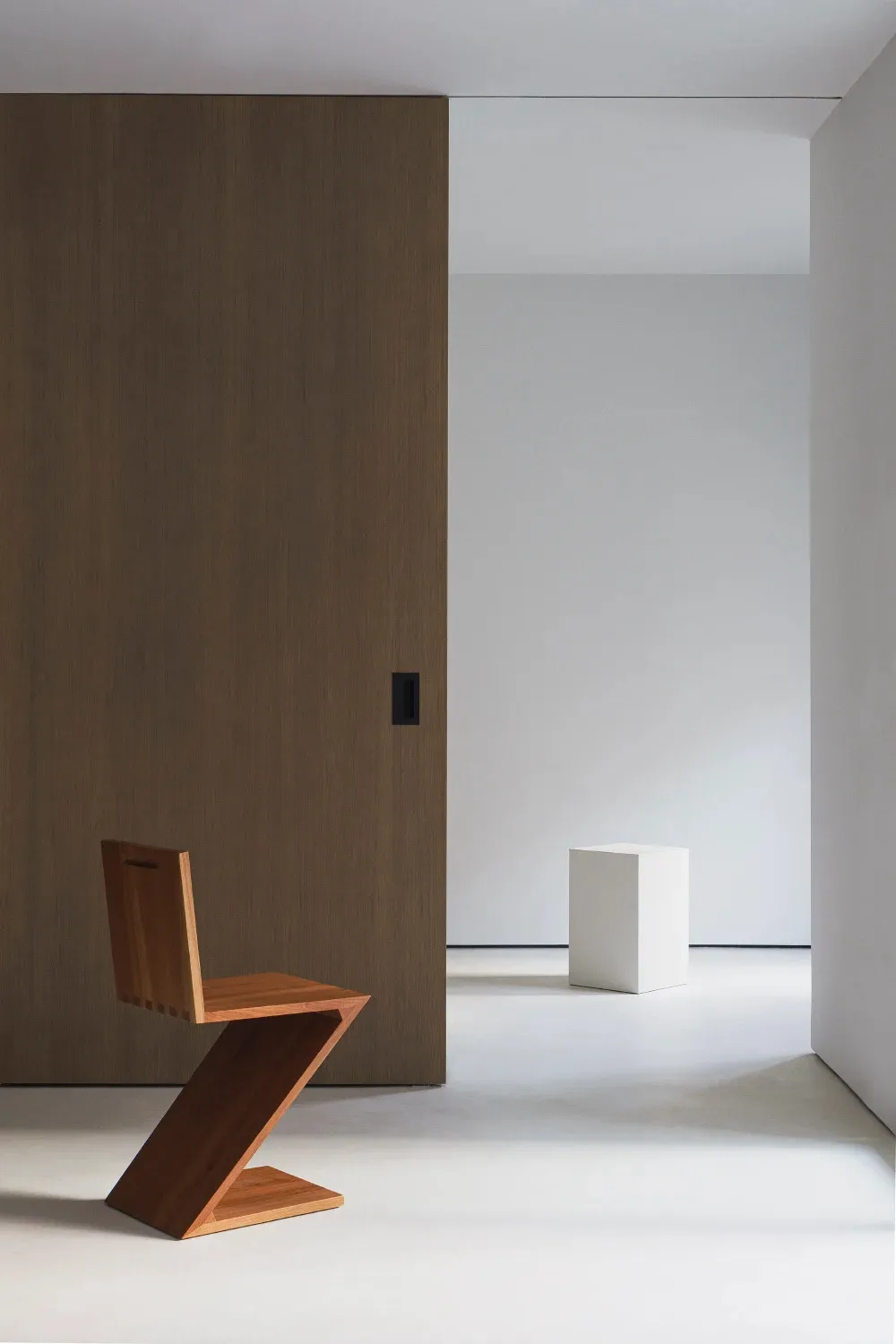 A minimalist space featuring a wooden zigzag chair and a white cube on a polished concrete floor.