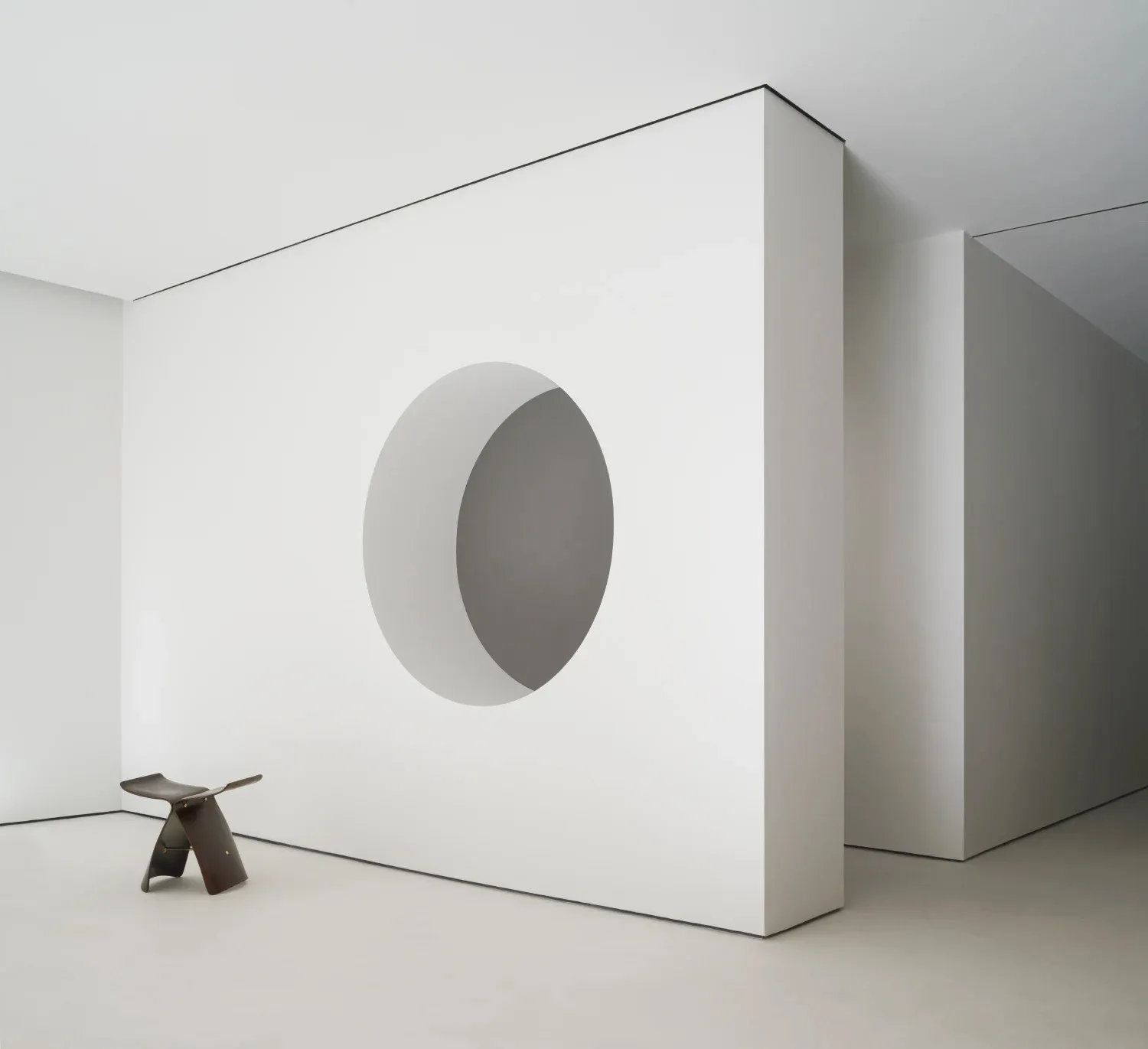 A minimalist white concrete room with a circular cutout in the wall and a wooden stool in a white interior setting.