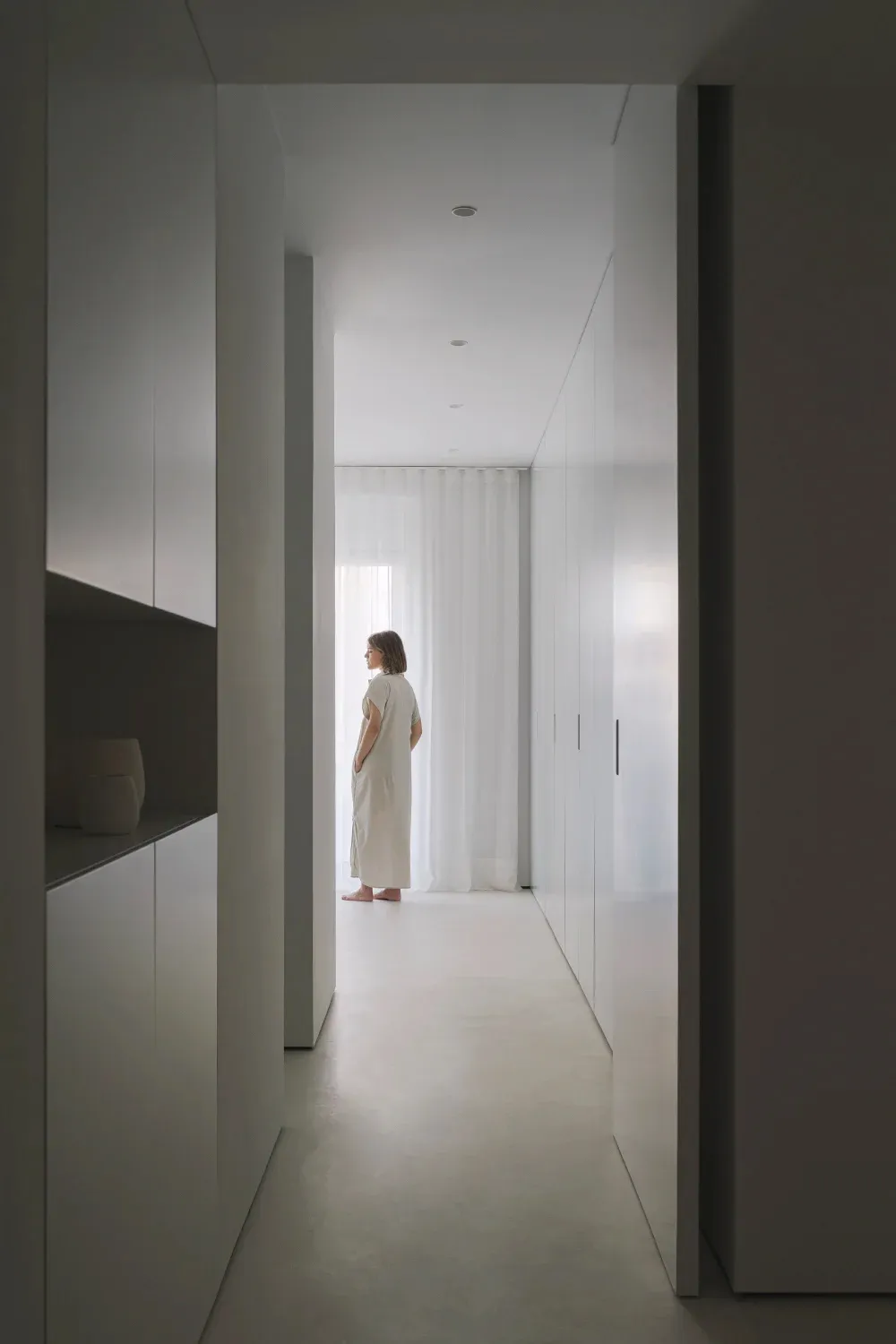 A minimalist hallway with white walls and flooring, featuring a person standing by a window.
