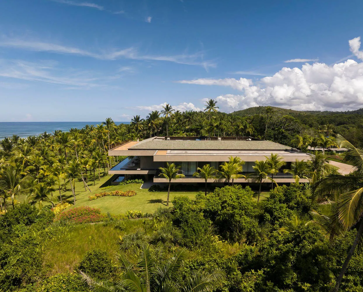 A modern beach house with concrete exterior materials surrounded by lush greenery and palm trees.