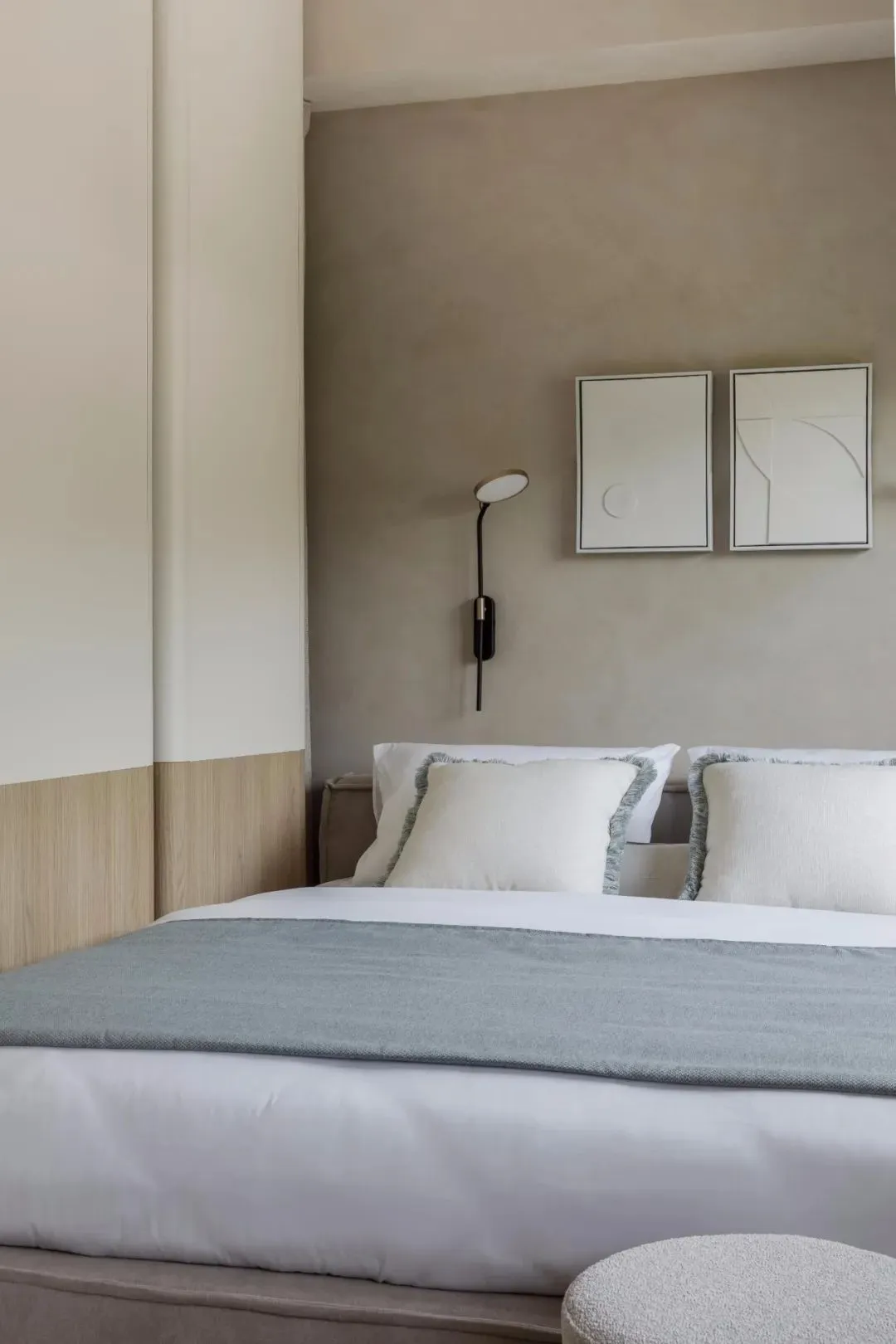 A minimalist bedroom with gray and white bedding, framed abstract art, and a modern fabric ottoman.
