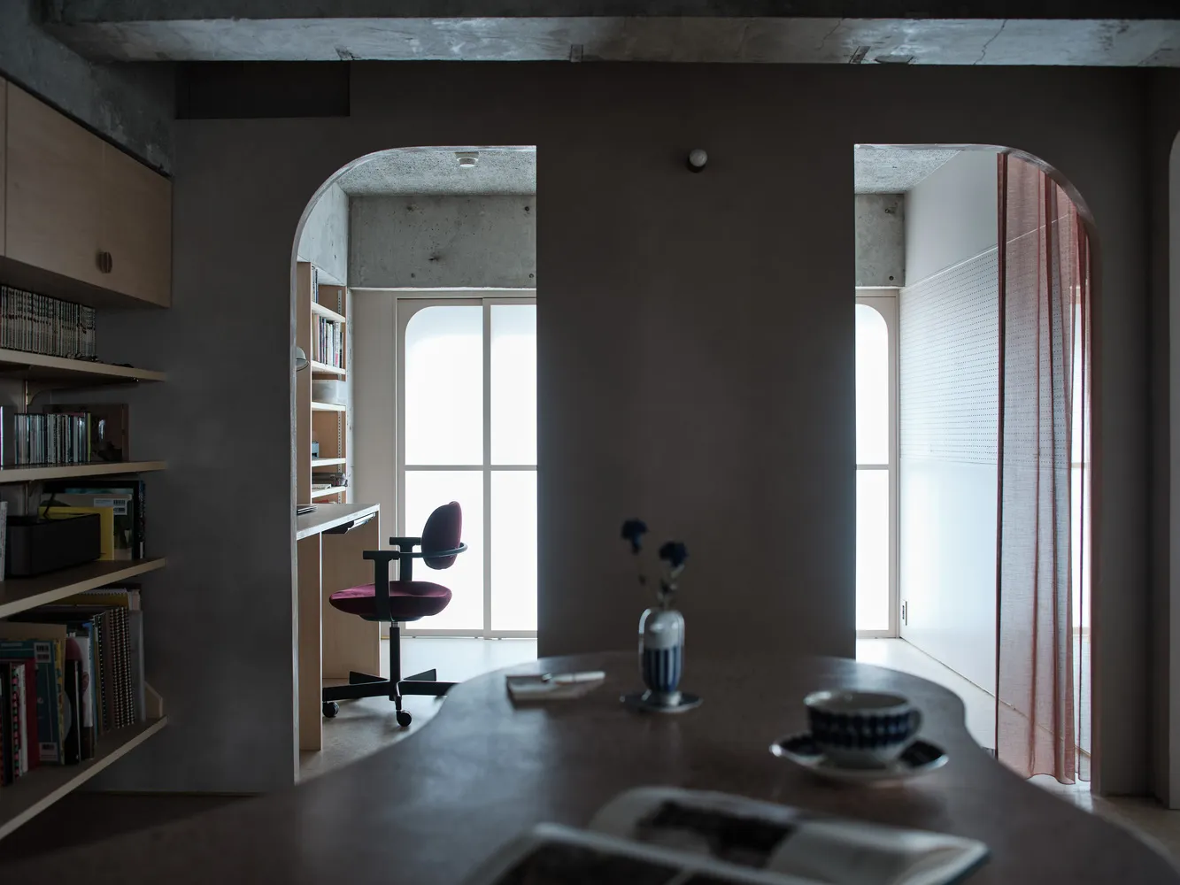 A modern minimalist home office features a concrete wall with an arched doorway leading to a wooden desk.