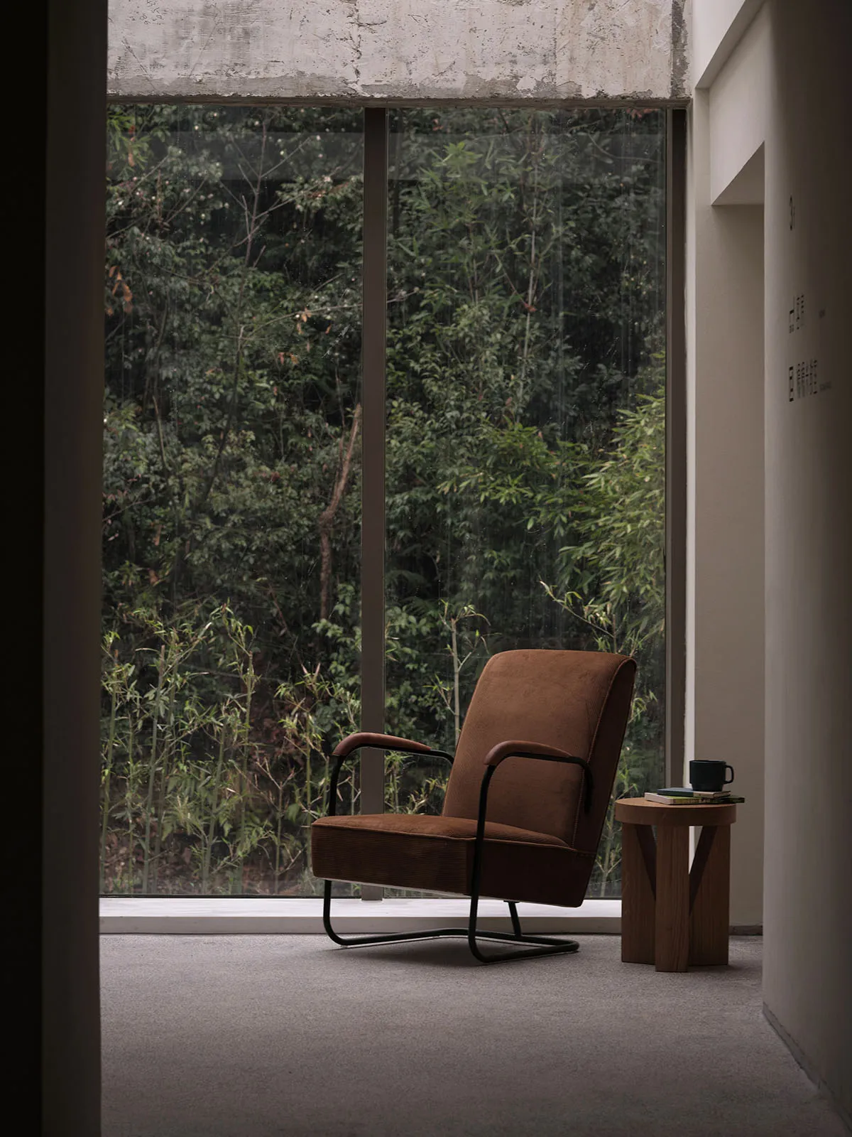 An industrial-style living room with a brown fabric armchair, metal frame, and glass window overlooking a forest.